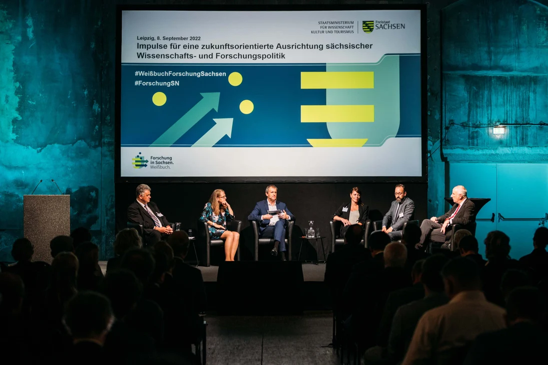Blick auf das Podium vor dem Screen »Impulse für eine zukunftsorientierte Ausrichtung sächsischer Wissenschafts- und Forschungspolitik«