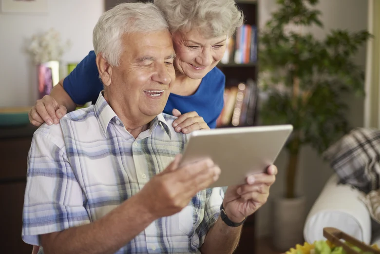 Grauhaariger Mann mit einem Tablet, dem eine grauhaarige Frau über die Schulter schaut.
