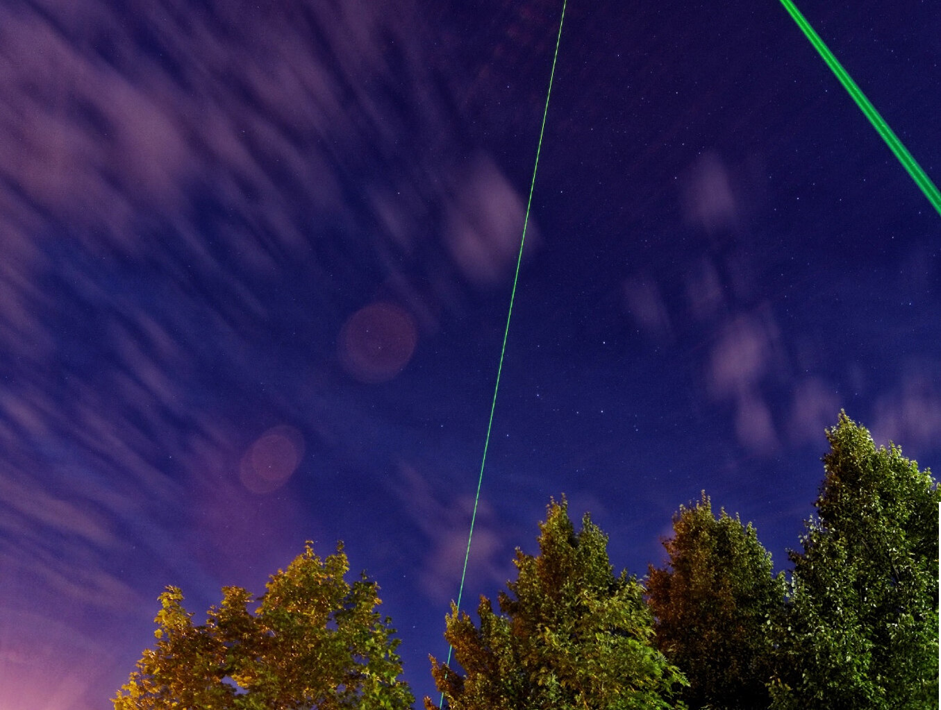 Drei grüne Laserstrahlen schießen in den violetten Himmel über Baumwipfel.