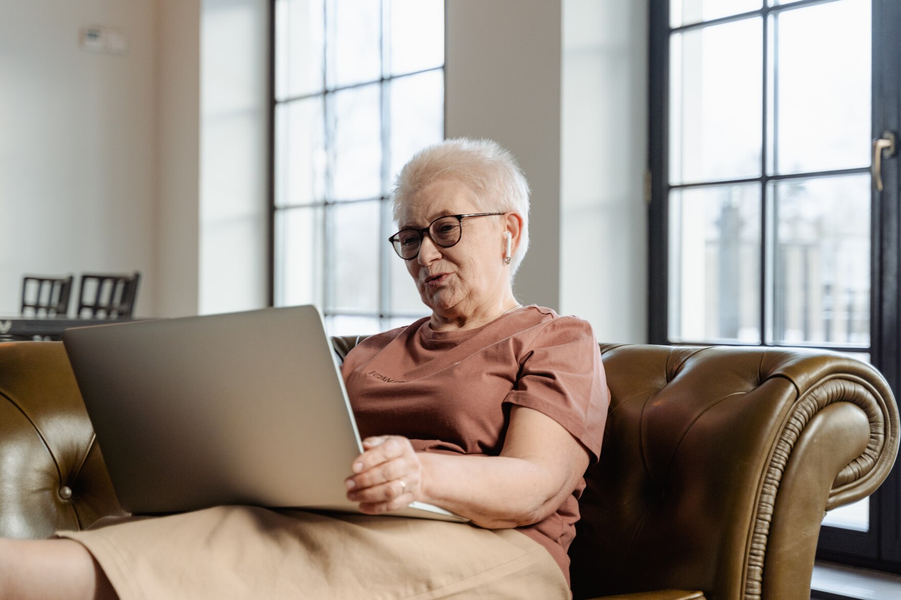 Ältere, grauhaarige Frau mit Brille, In-Ear-Kopfhörern und einem Laptop entspannt auf einem brauenen Ledersofa sitzend.